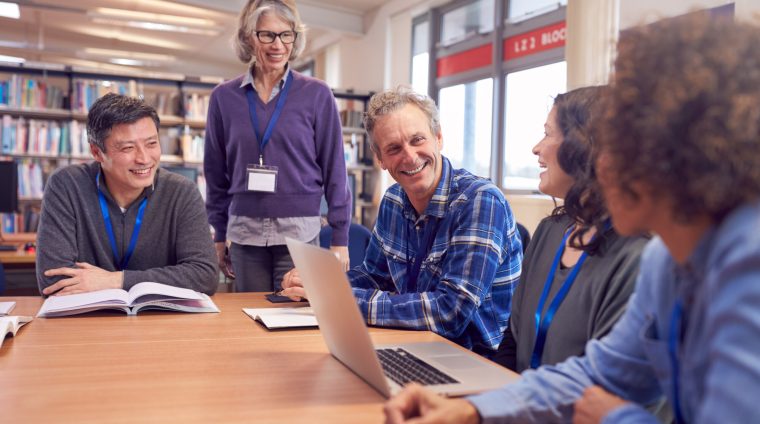 Teacher With Group Of Mature Adult Students In Class Sit Around Table And Work In College Library