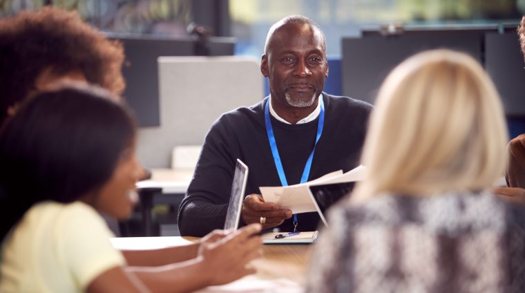 University Or College Students Sitting Around Table With Tutor Collaborating In Seminar On Laptop